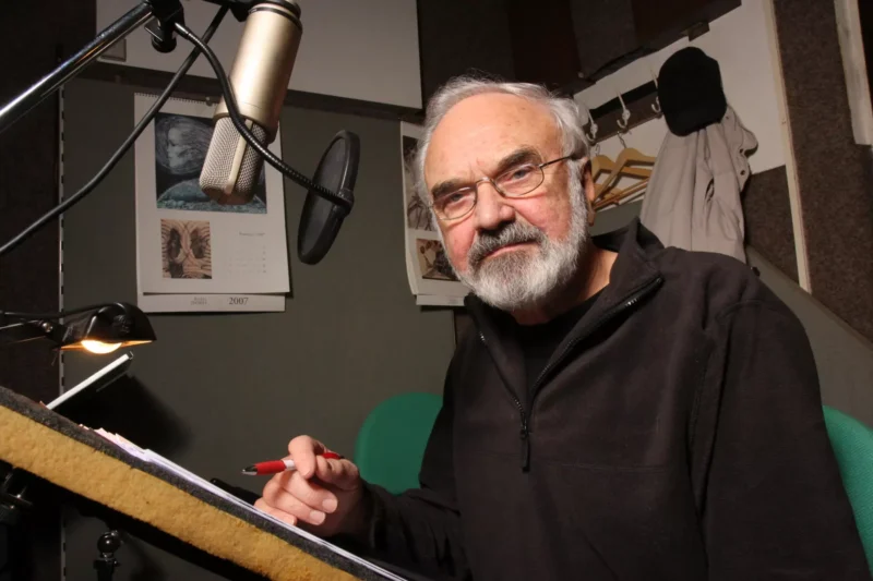 Starší muž s bradou a okuliarmi sedí v nahrávacom štúdiu, drží pero a papier, blízko mikrofónu. Prostredie je príjemné a sústredené.