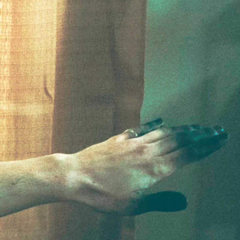 Ruka s prsteňom, čiastočne v tieni, sa tiahne oproti jemne osvetlenej oranžovej opone. Obraz vyvoláva tajomnú, introspektívnu náladu.