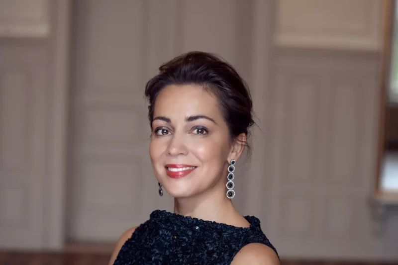 Žena sa usmieva vnútri, má na sebe čierne textúrované šaty a visiace náušnice. Má elegantný, sebavedomý výraz s neutrálne tónovanými stenami za sebou.