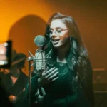Žena spieva do starožitného mikrofónu a vrúcne sa usmieva. Má na sebe okuliare a dlhé vlasy jej padajú cez plecia. Pódium má teplú, zlatistú žiaru.