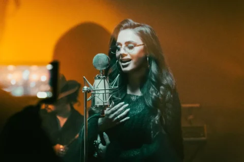 Žena spieva do starožitného mikrofónu a vrúcne sa usmieva. Má na sebe okuliare a dlhé vlasy jej padajú cez plecia. Pódium má teplú, zlatistú žiaru.