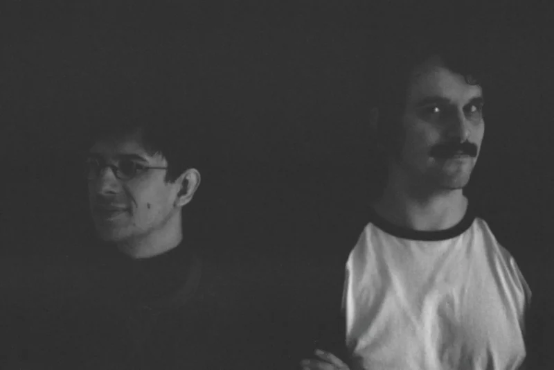 Čiernobiela fotografia dvoch mužov v slabo osvetlenom priestore. Jeden má okuliare a usmieva sa, zatiaľ čo druhý má fúzy a neutrálny výraz. Náladová atmosféra.