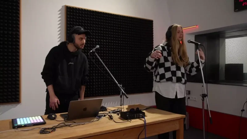 Dvaja hudobníci v štúdiu, jeden s notebookom a audio vybavením, druhý spieva do mikrofónu. Vyzerajú sústredene a majú kreatívnu atmosféru.