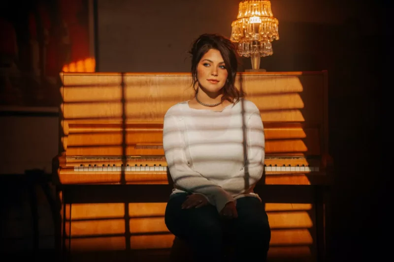 Žena sedí pred klavírom, osvetlená teplým, pruhovaným slnečným svetlom presvitajúcim cez žalúzie. Má na sebe biely sveter a modré džínsy, z ktorých vyžaruje pokojná, introspektívna nálada.