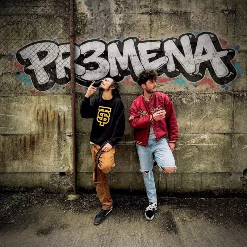 Dvaja muži stoja pri stene pokrytej graffiti s tučným nápisom „PR3MENA“. Jeden fajčí, obaja prejavujú ležérnu mestskú atmosféru.