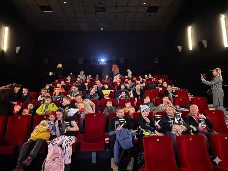 Preplnené kino s ľuďmi rôzneho veku sediacimi na červených stoličkách s pukancami a občerstvením v rukách. Atmosféra je živá a očakávaná.