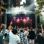 Dav na vonkajšom koncerte dvíha ruky blízko nápisu „REC. STAGE“. Jasné svetlá pódia žiaria uprostred industriálneho pozadia a dymovej atmosféry a napĺňajú scénu energiou.