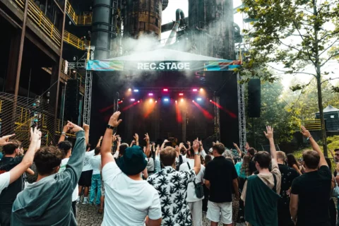 Dav na vonkajšom koncerte dvíha ruky blízko nápisu „REC. STAGE“. Jasné svetlá pódia žiaria uprostred industriálneho pozadia a dymovej atmosféry a napĺňajú scénu energiou.