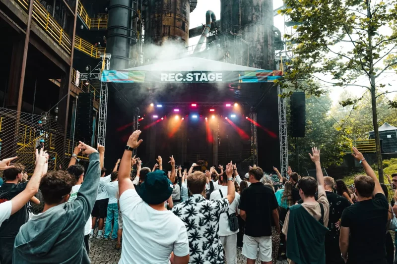Dav na vonkajšom koncerte dvíha ruky blízko nápisu „REC. STAGE“. Jasné svetlá pódia žiaria uprostred industriálneho pozadia a dymovej atmosféry a napĺňajú scénu energiou.