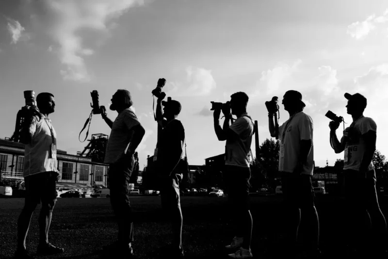 Siluety fotografov držia fotoaparáty smerom k oblohe počas západu slnka. Priemyselné budovy v pozadí pod čiastočne zamračenou oblohou. Nálada a umelecké cítenie.