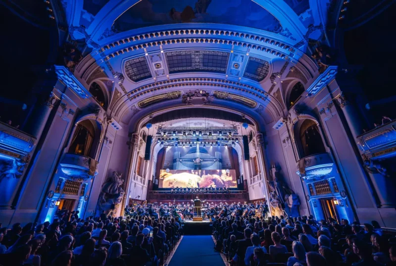 Veľkolepá koncertná sála so zdobenou architektúrou je zaliata modrým svetlom. Na pódiu vystupuje celý orchester s dirigentom v čele, sledovaný veľkým publikom.