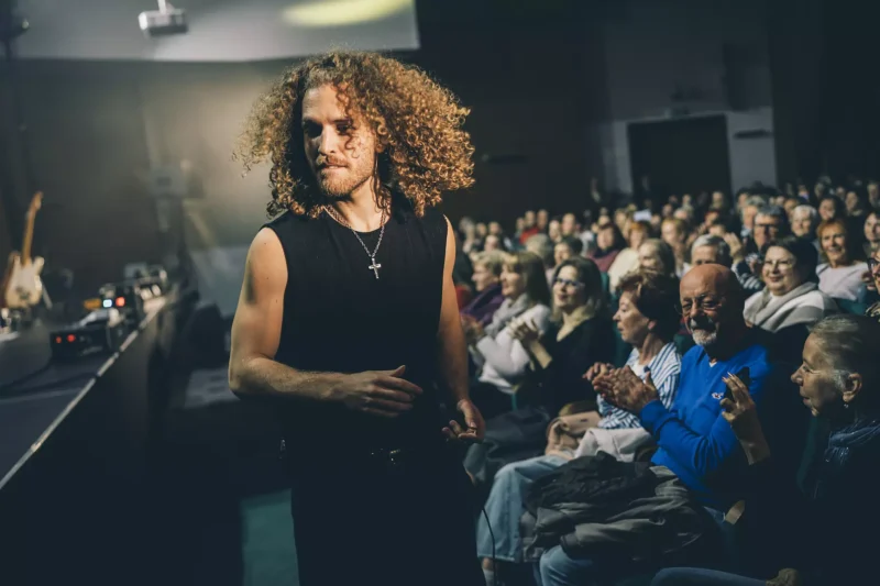 Muž s kučeravými vlasmi a krížikom v ruke prechádza cez tlieskajúce publikum v slabo osvetlenom priestore. Atmosféra je živá a pútavá.