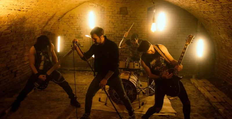 Rocková kapela intenzívne vystupuje v slabo osvetlenom podzemnom priestore s tehlovými stenami. Gitarista a spevák stoja vpredu a sršia vysokou energiou.