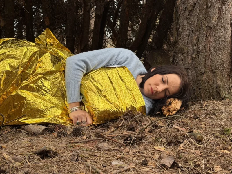 Žena spí vonku, zabalená v reflexnej žltej núdzovej deke, s hlavou opretou o poleno blízko stromu, čo vyvoláva pocit prežitia a pokoja.