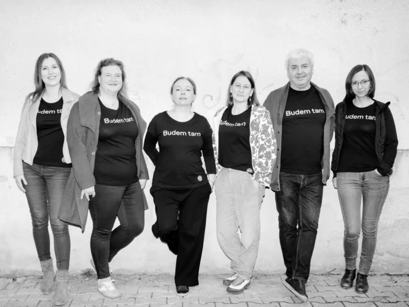 Skupina šiestich ľudí stojí vedľa seba pri obyčajnej stene v rovnakých tričkách s nápisom „Budem tam“. Vyzerajú uvoľnene a usmievajú sa. Čiernobiela fotografia.