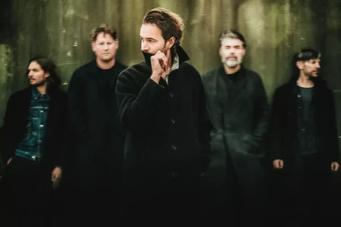 Skupina piatich mužov v tmavom oblečení stojí na rozmazanom, tlmenom pozadí. Centrálna postava hľadí do strany a vytvára tak zamyslenú atmosféru.