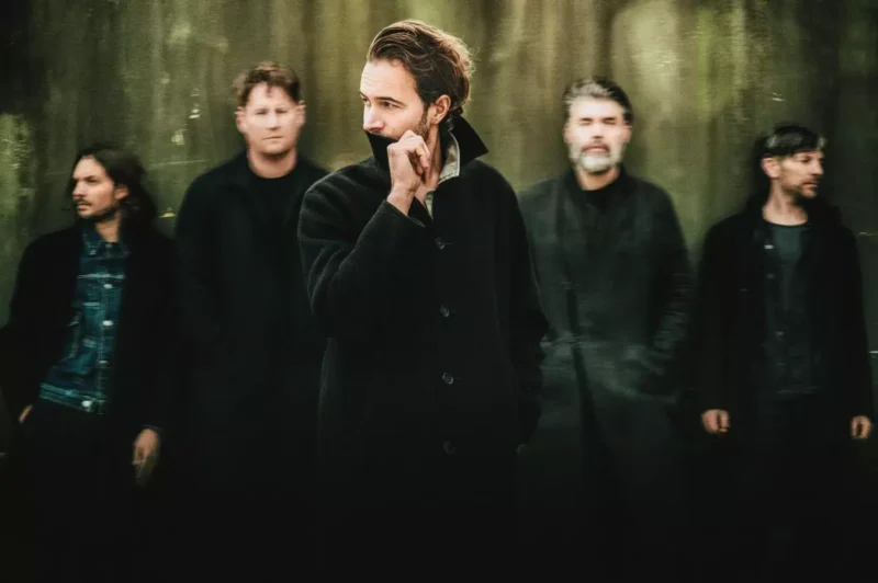 Skupina piatich mužov v tmavom oblečení stojí na rozmazanom, tlmenom pozadí. Centrálna postava hľadí do strany a vytvára tak zamyslenú atmosféru.