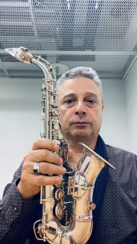 Muž drží saxofón v miestnosti so sieťovaným stropom. Má na sebe tmavú bodkovanú košeľu a sústredený výraz, čo naznačuje jeho oddanosť hudbe.