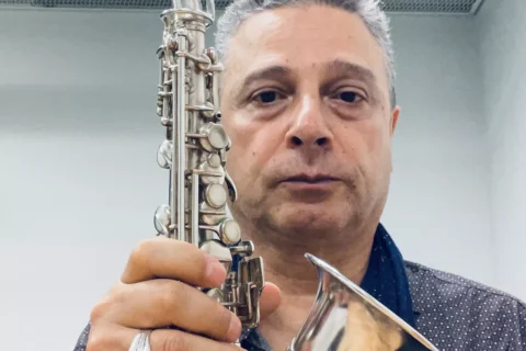 Muž drží saxofón v miestnosti so sieťovaným stropom. Má na sebe tmavú bodkovanú košeľu a sústredený výraz, čo naznačuje jeho oddanosť hudbe.