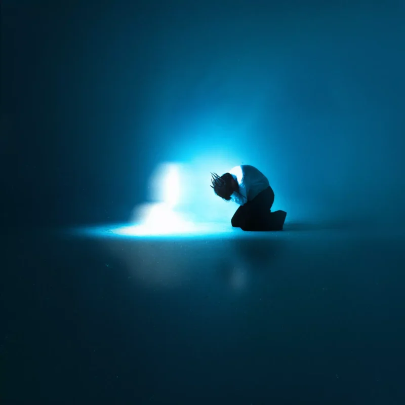 Človek kľačí na podlahe, zahalený v modrom svetle, ktoré evokuje pocit samoty a introspekcie. Tlmená, éterická žiara vytvára melancholickú atmosféru.