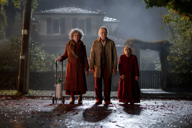 Traja starší ľudia stoja vážne na ulici v noci, s jesenným lístím na zemi a hmlistým domom v pozadí. Jeden používa kyslíkovú fľašu.