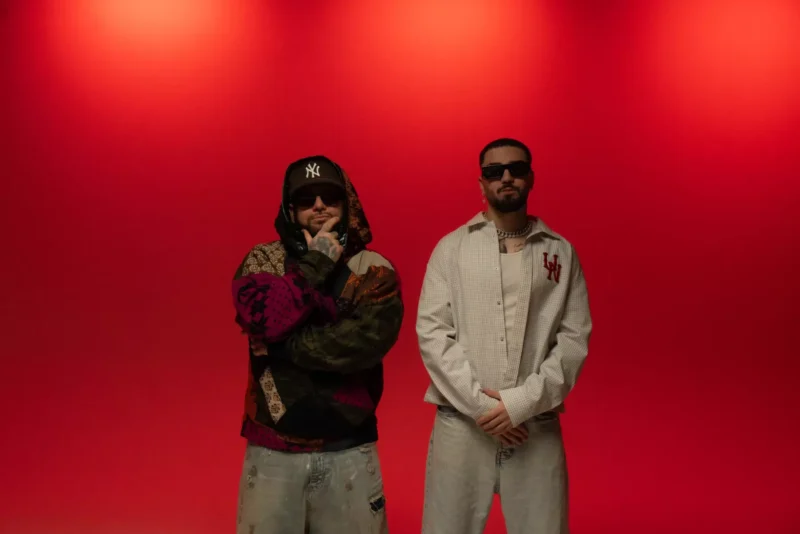 Dvaja muži stoja sebavedomo na červenom pozadí. Jeden má na sebe farebnú bundu a čiapku, druhý vzorovanú košeľu. Obaja majú slnečné okuliare.