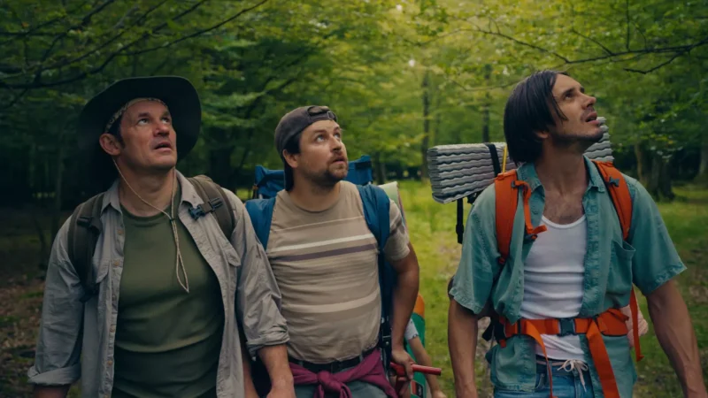 Traja muži s batohmi hľadia hore v lese a vyzerajú zvedavo. Bujná zeleň ich obklopuje dobrodružnou a objavovateľskou atmosférou.