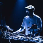 DJ s plochou šiltovkou a okuliarmi, osvetlený modrými svetlami pódia, sa sústredene sústredí na mixovanie hudby v DJskom pulte plnom káblov a vybavenia.