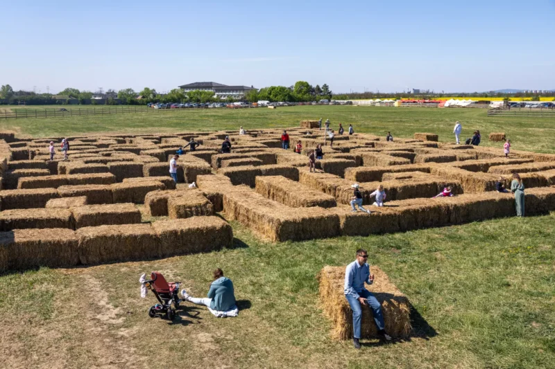 Ľudia si užívajú slnečný deň v malom bludisku z balíkov sena na trávnatej lúke. Niektorí sedia na balíkoch sena, zatiaľ čo iní skúmajú bludisko a vytvárajú tak uvoľnenú a hravú atmosféru.