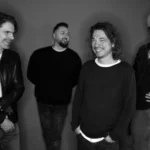 Čiernobiela fotografia štyroch mužov v uvoľnenej póze, usmievajúcich sa a smejúcich sa. Majú na sebe ležérne oblečenie, jeden z nich má klobúk a slnečné okuliare, čo vyjadruje radostný tón.