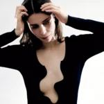 Žena v čiernom tope s hlbokým vlnitým výstrihom pózuje na čistom bielom pozadí. Jej výraz je pokojný, ruky má vo vlasoch.