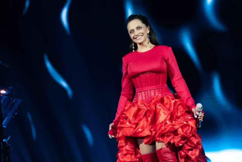 Žena v žiarivo červených šatách sebavedomo stojí na pódiu a drží mikrofón. Pozadie je doplnené jemným modrým osvetlením, ktoré vytvára dramatickú a živú atmosféru.