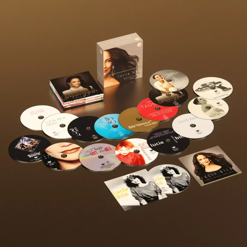 Na hnedom povrchu je rozložená zbierka CD s farebnými obalmi albumov a krabicou s portrétom ženy. Nálada je živá a oslavná.