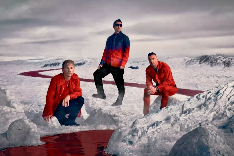 Traja muži v červených oblekoch stoja a kľačia na zľadovatenom teréne. Krajinou pod zamračenou oblohou preteká červená rieka, ktorá vytvára dramatickú scénu.