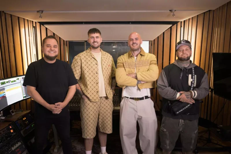 Štyria muži stoja s úsmevom v útulnom nahrávacom štúdiu s drevenými panelmi. Vyžarujú uvoľnenú a kolaboratívnu atmosféru, obklopení audiotechnikou.