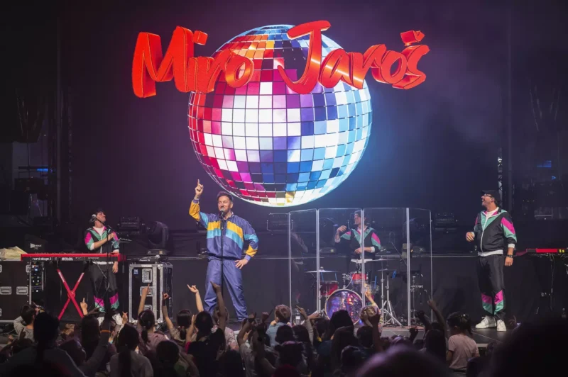 Na pódiu sú účinkujúci vo farebných teplákových súpravách, v strede je hudobník so zdvihnutým prstom. Za nimi svieti veľká disko guľa a nápis „Miro Jaroš“. V hľadisku stoja deti a vytvárajú energickú atmosféru.