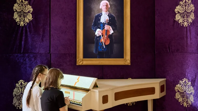 Dve deti hrajú na klávesoch pod portrétom historickej postavy s husľami v zlatom ráme na zdobenom fialovom pozadí.