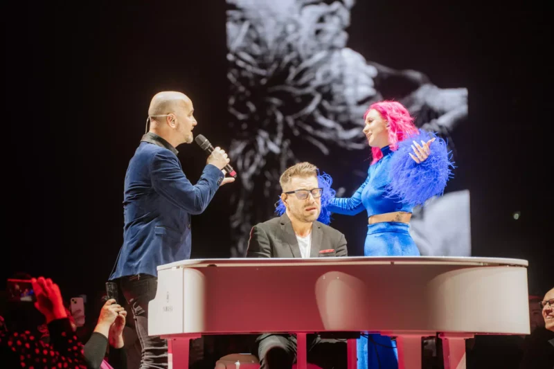 Na pódiu stoja traja účinkujúci s bielym klavírom. Jeden spieva do mikrofónu, druhý hrá na klavíri a tretí, v žiarivo modrom oblečení, výrazne gestikuluje.