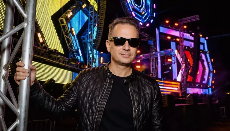 Muž v čiernej prešívanej bunde a slnečných okuliaroch sebavedomo stojí na vonkajšom koncerte, obklopený žiarivými, farebnými svetlami a konštrukciami pódia.