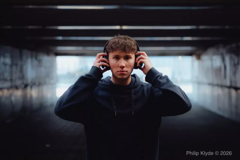 Mladý muž so slúchadlami pod slabo osvetleným nadjazdom hľadí priamo do kamery. Zdá sa byť sústredený, čo vytvára náladovú a introspektívnu atmosféru.