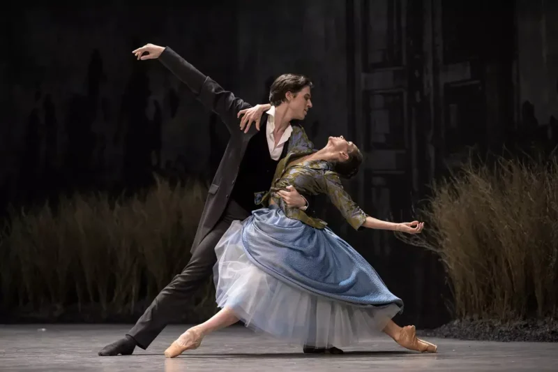 Baletný tanečník a tanečnica predvádzajú elegantnú pózu na javisku. Žena je v špičkách a má na sebe modré šaty. Pozadie je tmavé s vysokou trávou, čo vytvára dramatickú atmosféru.