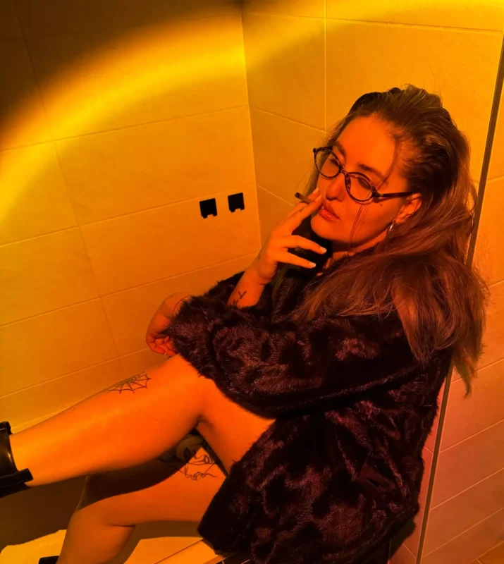 Dlhovlasý muž s okuliarmi a kožuchom zamyslene sedí v slabo osvetlenej miestnosti s teplým oranžovým svetlom a vyžaruje z nej uvoľnenú atmosféru.