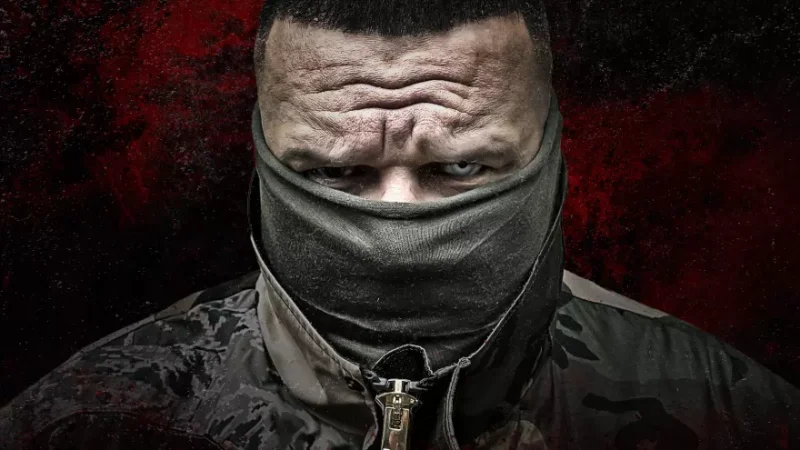 Osoba s napätým výrazom uprene hľadí, čiastočne zakrytá tmavou maskou a bundou, na drsnom červeno-čiernom pozadí.