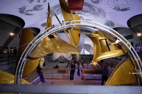 Futuristická umelecká inštalácia s veľkými zlatými štruktúrami a kovovými oblúkmi v priestrannej galérii. Ľudia pozorujú a na strop sa premietajú kozmické vzory.