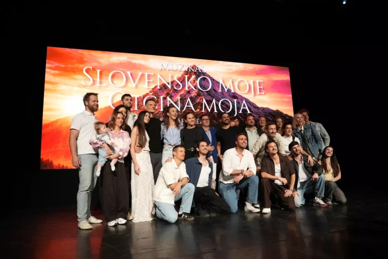 Skupina ľudí šťastne pózuje na pódiu pred žiarivým pozadím s nápisom „Slovensko Moje, Domovina Moja“. Vyzerajú radostne a oslavne.