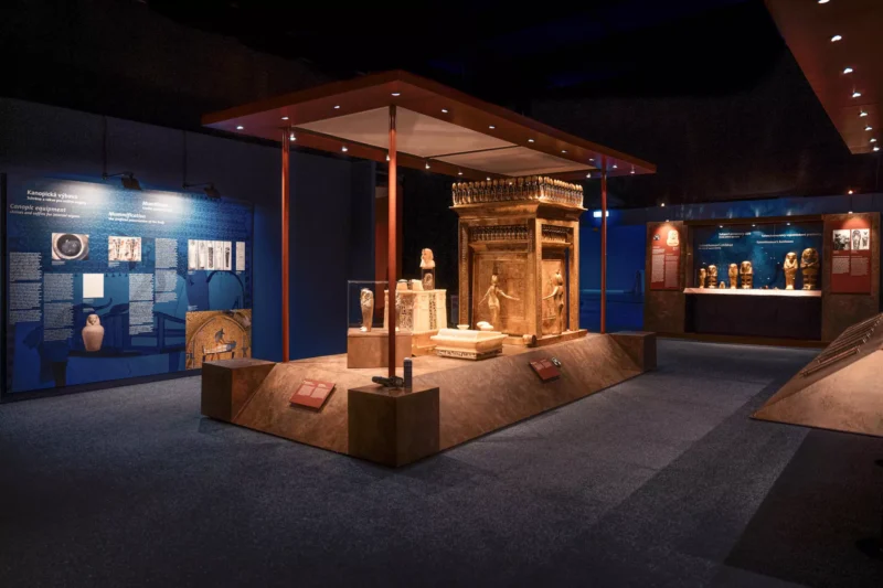 Staroveká egyptská výstavná miestnosť s veľkým, zdobeným sarkofágom v strede, obklopeným menšími artefaktmi a osvetlenými informačnými panelmi.