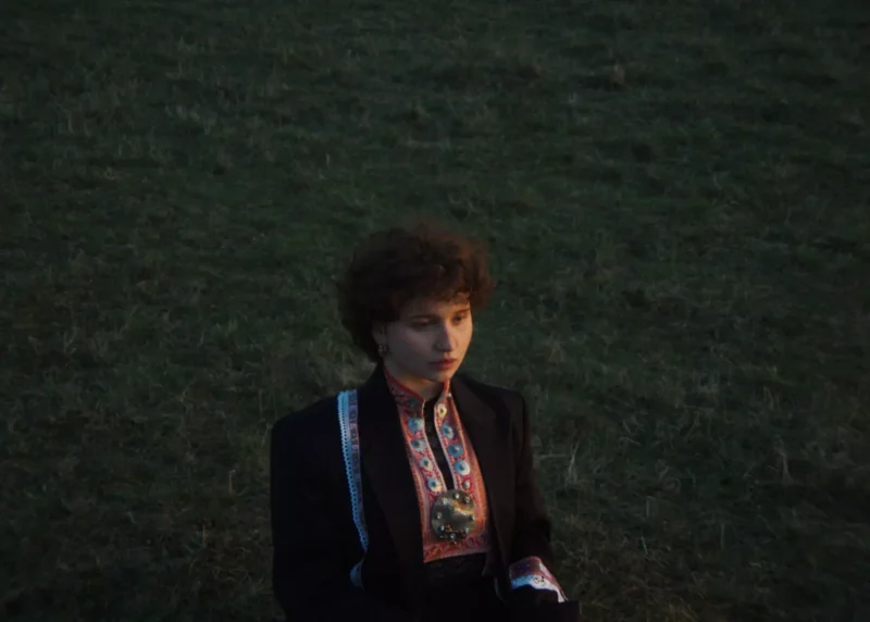 Muž s kučeravými vlasmi, oblečený v tradičnej vyšívanej bunde, zamyslene sedí na tráve za súmraku. Scéna vyvoláva pokojnú a introspektívnu náladu.