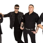 Štyria muži stoja sebavedomo na bielom pozadí, oblečení v čiernom oblečení. Jeden má slnečné okuliare, druhý má bielu kravatu. Všetci vyžarujú atmosféru rockovej kapely.