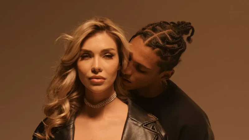 Žena s dlhými blond vlasmi a trblietavým chokerom sa sebavedomo pozerá dopredu. Muž s vrkočmi stojí za ňou a jemne sa k nej nakláňa. Obrázok má teplý, intímny tón.