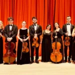 Sláčikový súbor vo formálnom čiernom oblečení pózuje so svojimi nástrojmi na pozadí žiarivo oranžovej opony a vyžaruje eleganciu a pohotovosť.